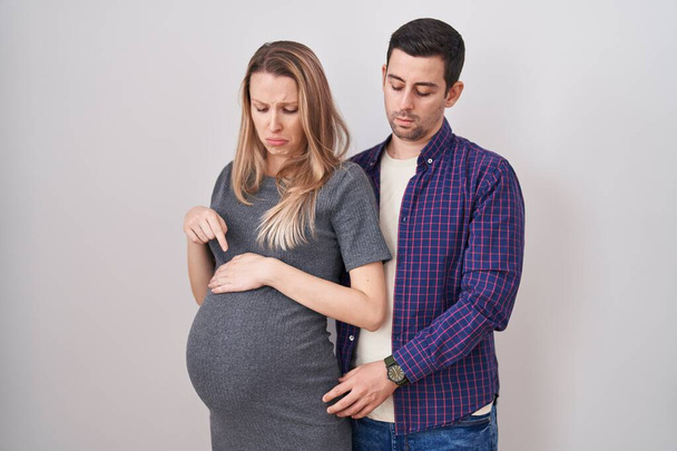 Junges Paar erwartet ein Baby, das vor weißem Hintergrund steht und traurig und aufgebracht nach unten zeigt, mit den Fingern die Richtung anzeigt, unglücklich und deprimiert.  - Foto, Bild