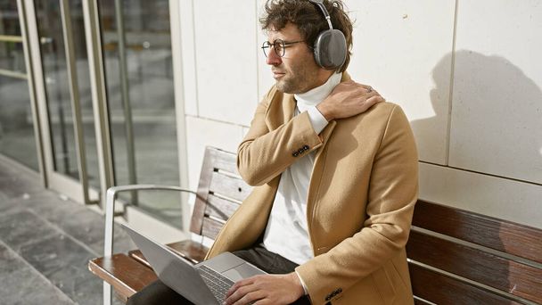 Hiszpan z brodą noszący słuchawki i dotykający szyi podczas pracy na laptopie siedzącym na ławce w miejskim otoczeniu. - Zdjęcie, obraz