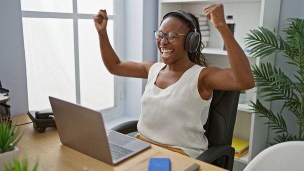Una mujer afroamericana adulta celebra con los brazos levantados en su lugar de trabajo, usando auriculares y gafas, con una computadora portátil y plantas de interior en el fondo. - Foto, Imagen