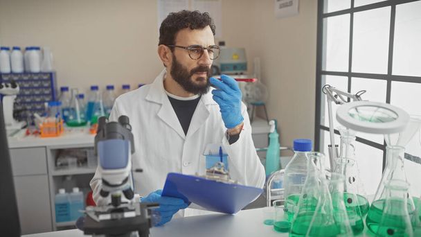 Ein bärtiger Mann mittleren Alters im Laborkittel untersucht ein Reagenzglas in einem gut ausgestatteten Labor mit Klemmbrett in der Hand. - Foto, Bild