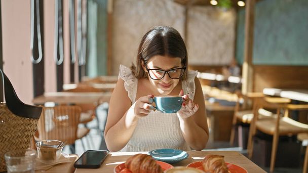 Χαρούμενη, όμορφη Ισπανίδα γυναίκα που απολαμβάνει ένα φλιτζάνι ζεστό καφέ γεμάτο καφεΐνη σε ένα ηλιόλουστο καφέ, το χαμόγελό της λάμπει καθώς χαλαρώνει σε ένα εσωτερικό χώρο σε ένα τραπέζι, γυαλιά λάμπει - Φωτογραφία, εικόνα