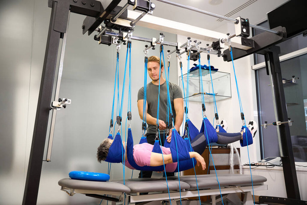 Young european male trainer helping middle aged woman hanging on suspension medical ropes after injury during recovery in rehabilitation center. Concept of modern healthy lifestyle - Photo, Image