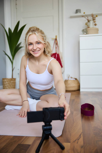Porträt einer jungen Frau als persönliche Yogalehrerin, die zu Hause Workout-Videos aufzeichnet, ihre Übungen mit der Digitalkamera protokolliert und Gummimatten benutzt. - Foto, Bild