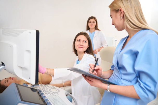Female gynecologist doing ultrasound scanning of pregnant belly of young woman while her colleague writing result of this examining and doctors looking at each other in clinic. Pregnancy and maternity - Photo, Image