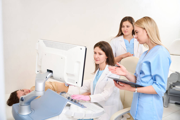 Female caucasian gynecologist doing ultrasound scanning of pregnant belly of young european woman while her colleague writing result of this examining in clinic. Concept of pregnancy and maternity - Photo, Image