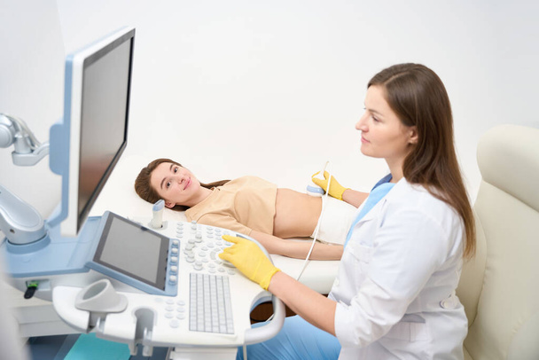 Vrouwelijke Kaukasische gynaecoloog doet echografie scannen van zwangere buik van jonge Europese vrouw in de moderne kliniek. Begrip zwangerschap en moederschap - Foto, afbeelding