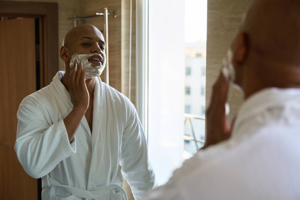 Jovem afro-americano manchando espuma de barbear no rosto e olhando para si mesmo no espelho no banheiro na hora da manhã. Conceito de procedimentos matinais e higiene - Foto, Imagem