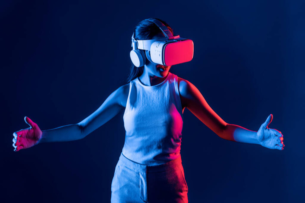 Умная женщина, стоящая в окружении неонового света, изнашивает VR-гарнитуру, соединяющую метавселенную, футуристическую технологию сообщества киберпространства, используя обе руки, взаимодействующие с генерируемым виртуальным объектом. Галлюцинация. - Фото, изображение