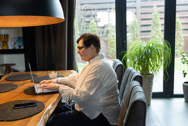 Взрослая пожилая женщина смотрит бизнес-обучение, онлайн-вебинар на ноутбуке компьютер удаленной работы или социального дистанционного обучения от дома. Видеоконференция для деловых женщин 60-х годов в виртуальном чате - Фото, изображение