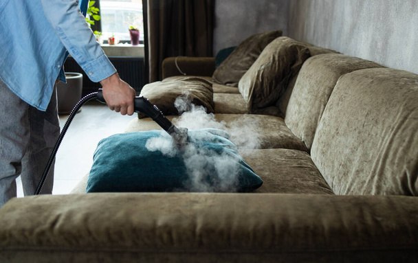 Ev temizliği. Bir adam kanepeyi ve mobilyaları buhar jeneratörüyle temizliyor. Dairenin temizlik servisi. Düz Los. Güçlü buhar basıncı. - Fotoğraf, Görsel