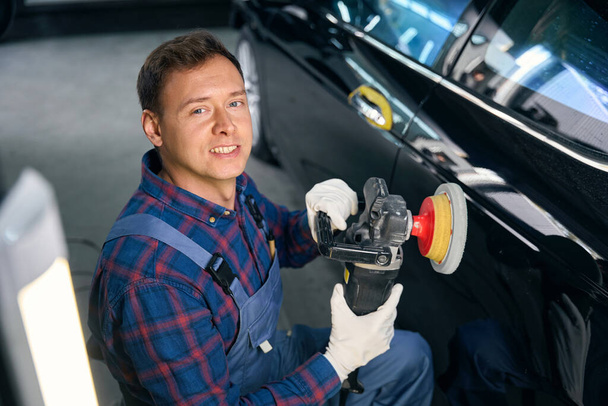 Mies tasaisessa ruumassa, kiillottaa auton yksityiskohtia, arvioi konepellin pinnan vaurioita. Pintakäsittely ennen maalausta - Valokuva, kuva