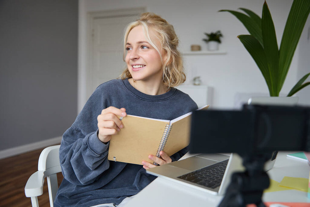 Porträt einer lächelnden blonden Frau, die im Schlafzimmer sitzt, Laptop und Digitalkamera benutzt, Videos für Lifestyleblog aufnimmt, liest, ihr Notizbuch benutzt. - Foto, Bild