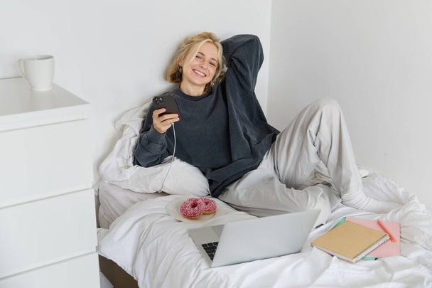 Portret van een jonge, mooie blonde vrouw, lui gevoel thuis, in bed liggen met een kopje thee, donut en laptop, op afstand studeren of online films kijken. - Foto, afbeelding