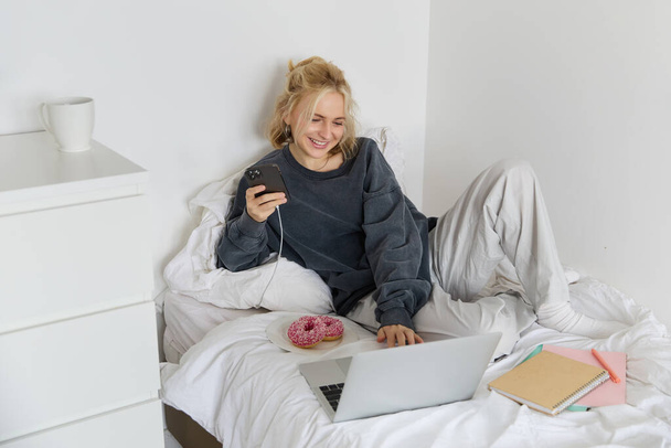 Lifestyle en mensen concept. Jonge mooie vrouw, thuisblijven, in bed liggen met laptop en smartphone, donut eten, genieten van vrije tijd, weekend thuis doorbrengen, film online kijken. - Foto, afbeelding