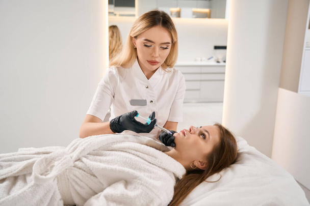 Skoncentrowany europejski kosmetolog wykonujący wstrzyknięcie kosmetyczne twarzy ze strzykawką młodej białej kobiety na kanapie w niewyraźnym salonie piękności. Pielęgnacja twarzy, podnoszenie i odmładzanie - Zdjęcie, obraz