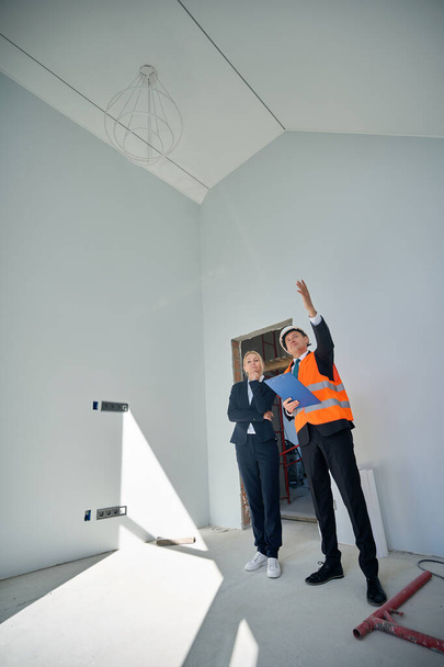 Teljes hosszúságú fénykép egy női ügyfélről és egy férfi építészről, amint egy befejezetlen szobában állnak, miközben ellenőrzik. - Fotó, kép