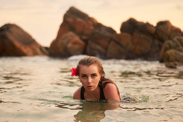 Female enjoys tranquil swim in nature setting, reflecting travel leisure. Woman with red flower in hair swims at dusk in serene rocky lagoon. Tourism adventure in natural sea pool, beauty moment. - 写真・画像