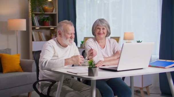 komunikacja online, radośni starsi rodzice wykorzystują rozmowy wideo z laptopami, aby komunikować się z rodziną za pomocą połączenia wideo, siedząc przy stole w pokoju - Materiał filmowy, wideo