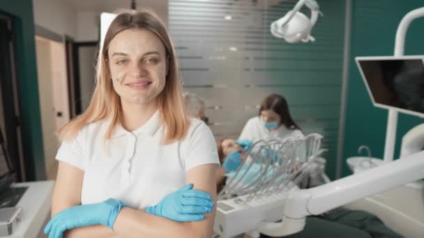 Z wyrazem zaufania, dentysta stoi przed kamerą, demonstrując swoją profesjonalną wiedzę, podczas gdy w tle inni lekarze pracują w celu zapewnienia wysokiej jakości leczenia stomatologicznego - Materiał filmowy, wideo