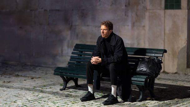 Мужчина сидит на скамейке в темноте. На нём чёрное пальто и рюкзак на коленях. Сцена тихая и мирная, мужчина смотрит вдаль - Фото, изображение