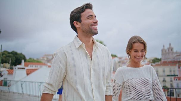 Romantik yeni evliler şehir manzarası olan terasta yürüyorlar. Yakışıklı esmer beyefendi Akdeniz 'de gülümseyen kız arkadaş elini öpüyor. Rahatlamış mutlu çift birlikte gülerken manzarayı seyrediyor. - Fotoğraf, Görsel