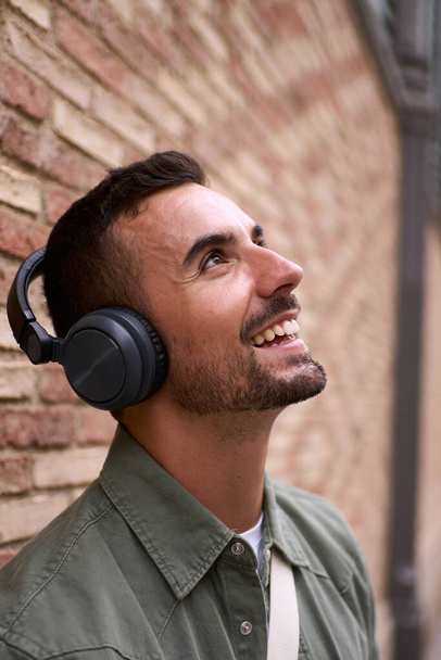 Vertical close up of handsome young adult tourist man looking up smiling with wireless headphones leaning against brick wall. His gesture suggests joy as he happy enjoys watching something outdoors - Фото, изображение