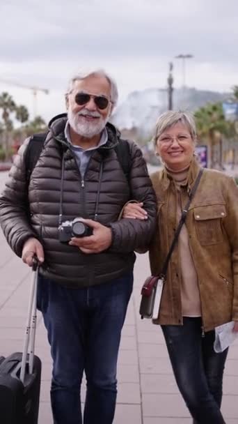 Vertikální fotoaparát přibližující usmívající se kavkazský dospělý turistický pár stojící pózující na ulici se zavazadly. Manžel a manželka užívají své důchodce prázdniny na sobě zimní oblečení na slunný den - Záběry, video