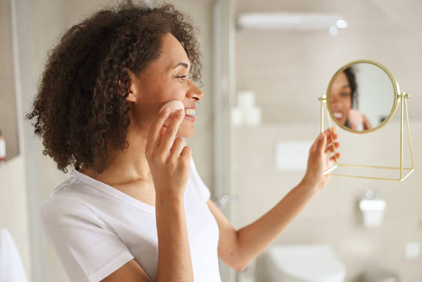 Uma mulher com uma toalha enrolada em torno dela está se admirando no espelho do banheiro. Ela está sorrindo e tocando seu rosto, pescoço e braço, enquanto gesticulando em direção a sua perna e coxa - Foto, Imagem