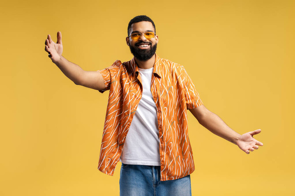 Vrolijk lachende Afro-Amerikaanse man in stijlvolle casual kleding en zonnebril met open armen kijkend naar camera geïsoleerd op gele achtergrond. Welkomstconcept - Foto, afbeelding