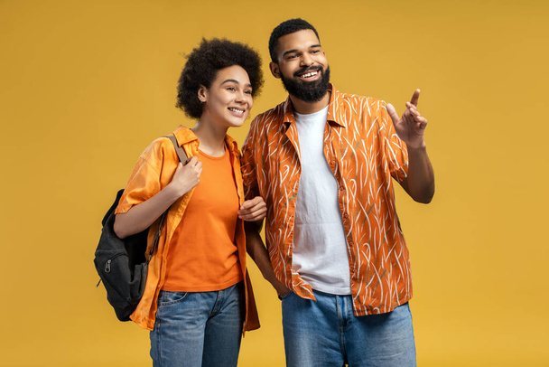 Glimlachende Afro-Amerikaanse man en vrouw met reisrugzak, vriendje met baard die iets naar zijn vriendin wees terwijl ze wegkeek, geïsoleerd op een gele achtergrond. Reisconcept - Foto, afbeelding