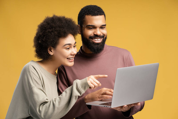 Attractive smiling African American couple, beautiful woman and man holding computer, using laptop, choosing, paying, watching video standing isolated on yellow background. Online shopping concept - Photo, Image