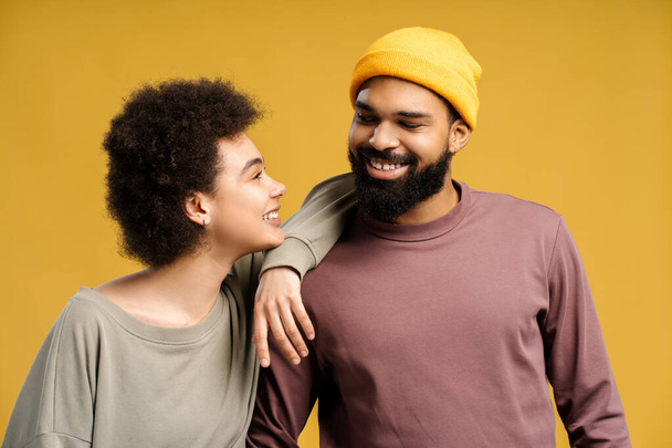 Attraente afroamericano barbuto uomo e donna con i capelli ricci, indossa vestiti alla moda, cappello giallo, abbracci parlando, in piedi isolato su sfondo giallo. Concetto di amore, comunicazione - Foto, immagini