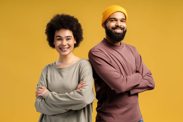 Cheerful, smiling African American couple, man wearing stylish yellow hat, happy woman with curly hair, with crossed arms standing isolated on yellow background. Relationship concept - Foto, imagen