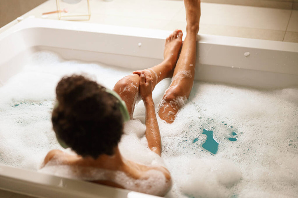 W łazience, kobieta wygodnie kąpie się w wannie wypełnionej bąbelkami z dzieckiem. Ich nogi są splecione, spoczywają na ciepłej drewnianej podłodze. - Zdjęcie, obraz