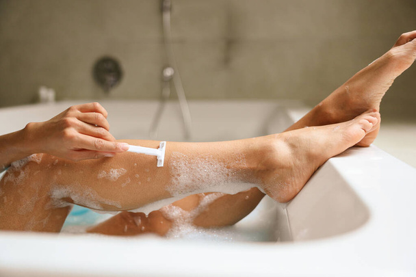 Μια γυναίκα ξυρίζει με χάρη τα πόδια της ενώ μουλιάζει σε ένα χαλαρωτικό μπάνιο, περιτριγυρισμένη από τη ζεστή αγκαλιά του ξύλου και του νερού - Φωτογραφία, εικόνα