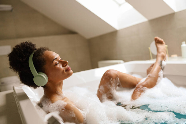 Een vrouw baadt ontspannen in een badkuip met een koptelefoon op in haar badkamer. Het water is tot aan haar borst, het creëren van een ontspannen en vredige gebeurtenis - Foto, afbeelding