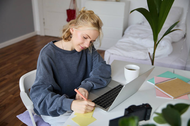 Portrait of smiling young woman, female student, doing distance learning course, using laptop, studying at home, writing down, making notes. - Фото, изображение