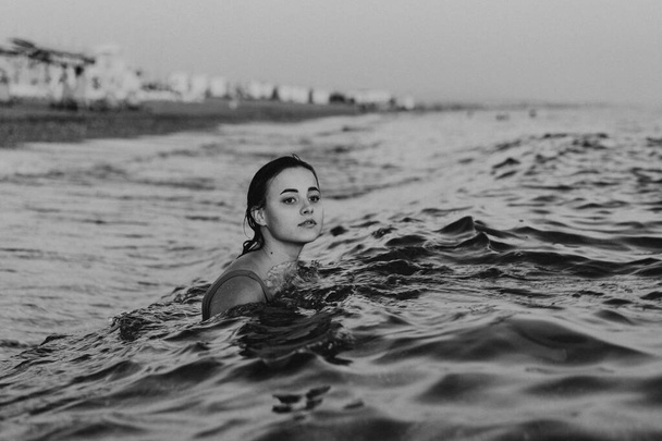 Een jonge vrouw met een glimlach op haar gezicht zwemt 's avonds vroeg in het rustige oceaanwater. Haar hoofd en schouders zijn zichtbaar boven de waterlijn - Foto, afbeelding