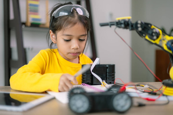 Egy fiatal lány ül egy asztalnál egy robot játékkal előtte. Egy jegyzetfüzetbe ír, valószínűleg a robotról, vagy a gondolatairól. A kíváncsiság és a kreativitás fogalma - Fotó, kép
