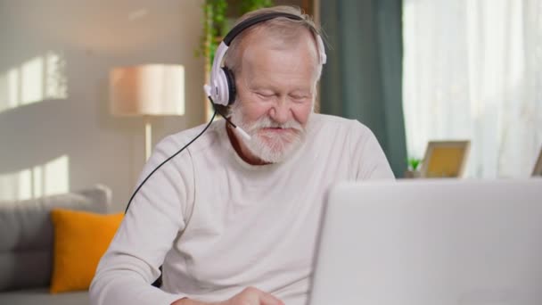 Современный пенсионер с седой бородой разговаривает на видео-вызове на ноутбуке с помощью гарнитуры и делает заметки в блокноте, сидя за столом в уютной комнате - Кадры, видео