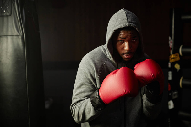 Jeune sportif afro-américain fort en gants de boxe rouges et sweat à capuche gris regardant la caméra ou le sac de boxe et se préparant pour le coup de pied - Photo, image