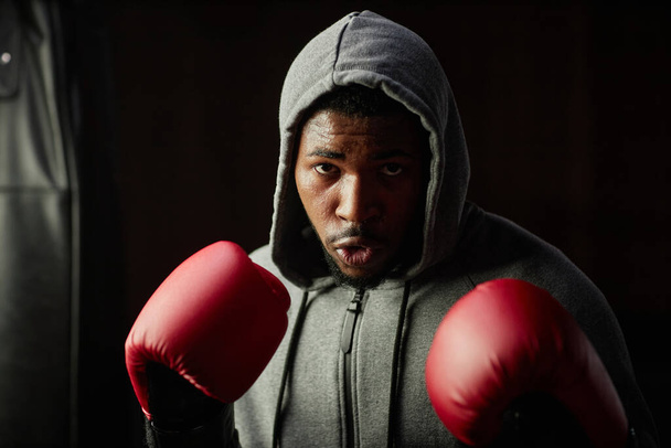 Retrato de un joven negro serio con capucha gris y guantes de boxeo rojos mirando a su rival durante el entrenamiento en el gimnasio o club deportivo - Foto, imagen