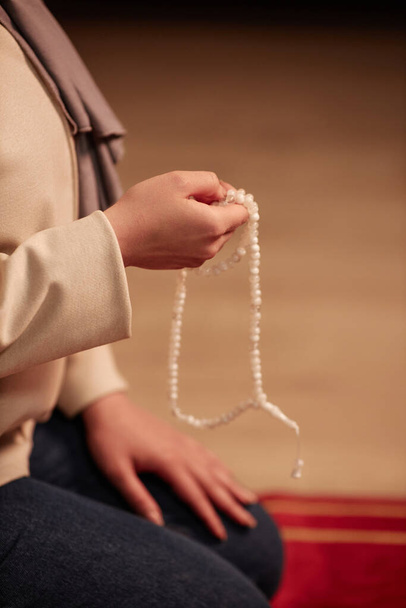 Nahaufnahme der Hand einer jungen islamischen Frau mit weißem Rosenkranz, die zu Hause vor der Kamera kniet und schweigend betet - Foto, Bild