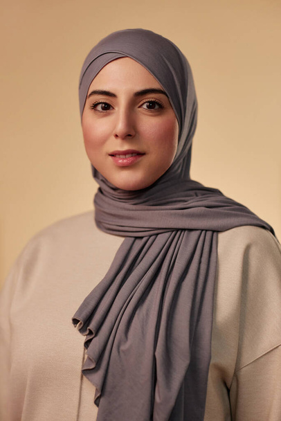 Jovem mulher muçulmana bonita em lenço de cabeça cinza e pulôver bege olhando para a câmera enquanto estava em isolamento contra o fundo pastel - Foto, Imagem