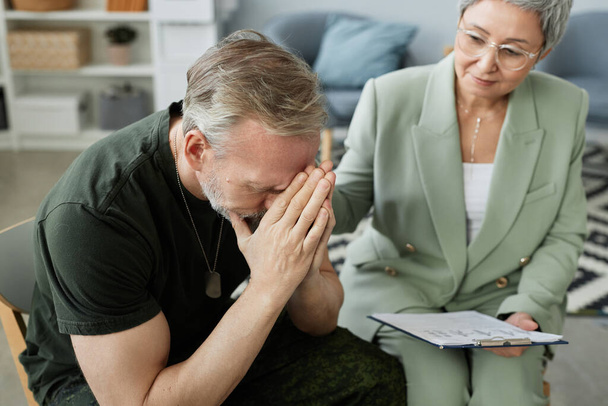 mature man with post traumatic syndrome keeping his hands put together close to face while sitting next to counselor supporting him at session - Photo, Image