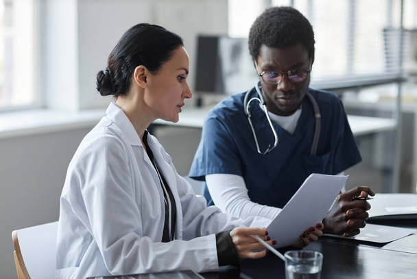 Clínica femenina confiada mostrando documento médico a un joven asistente masculino afroamericano en uniforme azul y explicándole los datos - Foto, Imagen