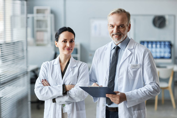 Dwóch doświadczonych lekarzy ogólnych w fartuchach laboratoryjnych patrzących przed kamerą w gabinecie medycznym, podczas gdy jeden z nich robi notatki w dokumencie - Zdjęcie, obraz