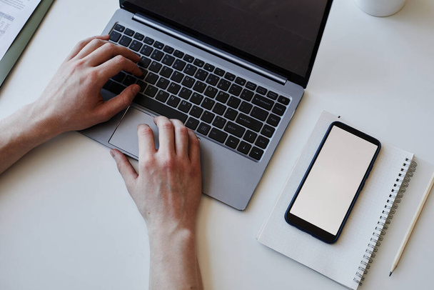 Вид сверху на человека, использующего ноутбук на столе в офисе, руки с макетом экрана смартфона на столе, копировальное пространство - Фото, изображение