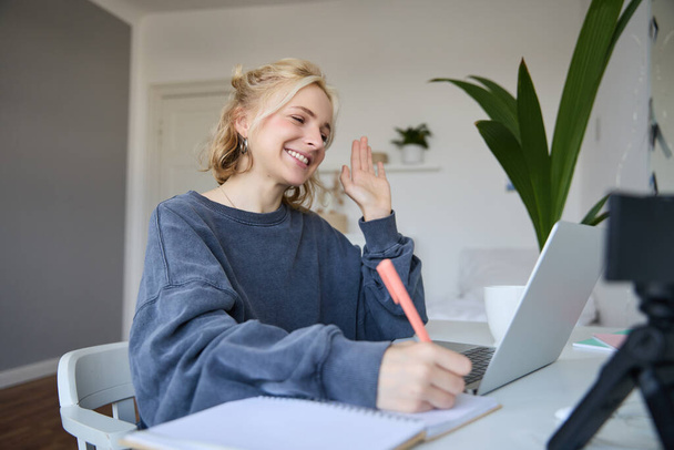 Portrait of young blond smiling woman, studying at home, remote education concept, connects to online course or lesson, writing in notebook. - Photo, Image