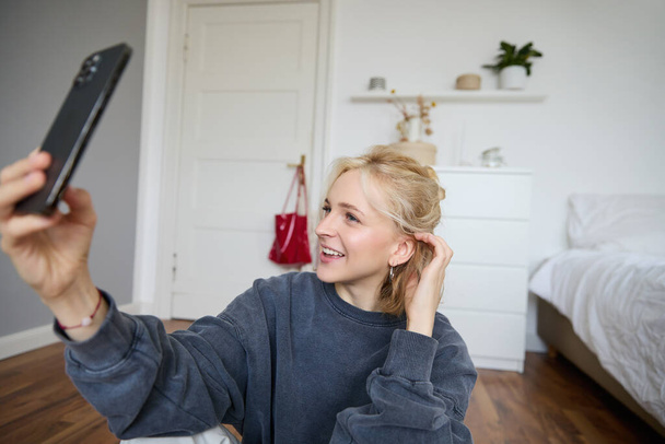 Retrato de una mujer joven, influyente de las redes sociales, tomando selfies en su habitación, sentada en el suelo, sosteniendo el teléfono inteligente y posando para una foto. - Foto, Imagen